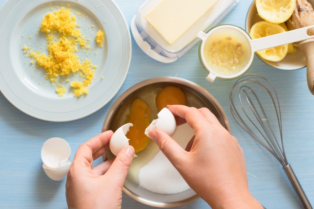 Woman cracking eggs into a bowl next to a block of butter and pile of lemon zest