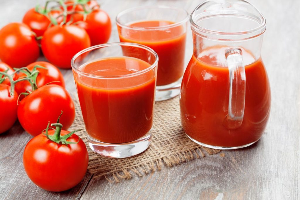 Glasses of tomato juice next to a pitcher of tomato juice with whole tomatoes on the side