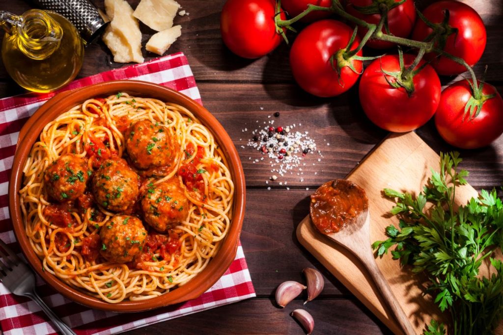 Bowl of spaghetti topped with meatballs and marinara sauce surrounded by tomatoes and herbs