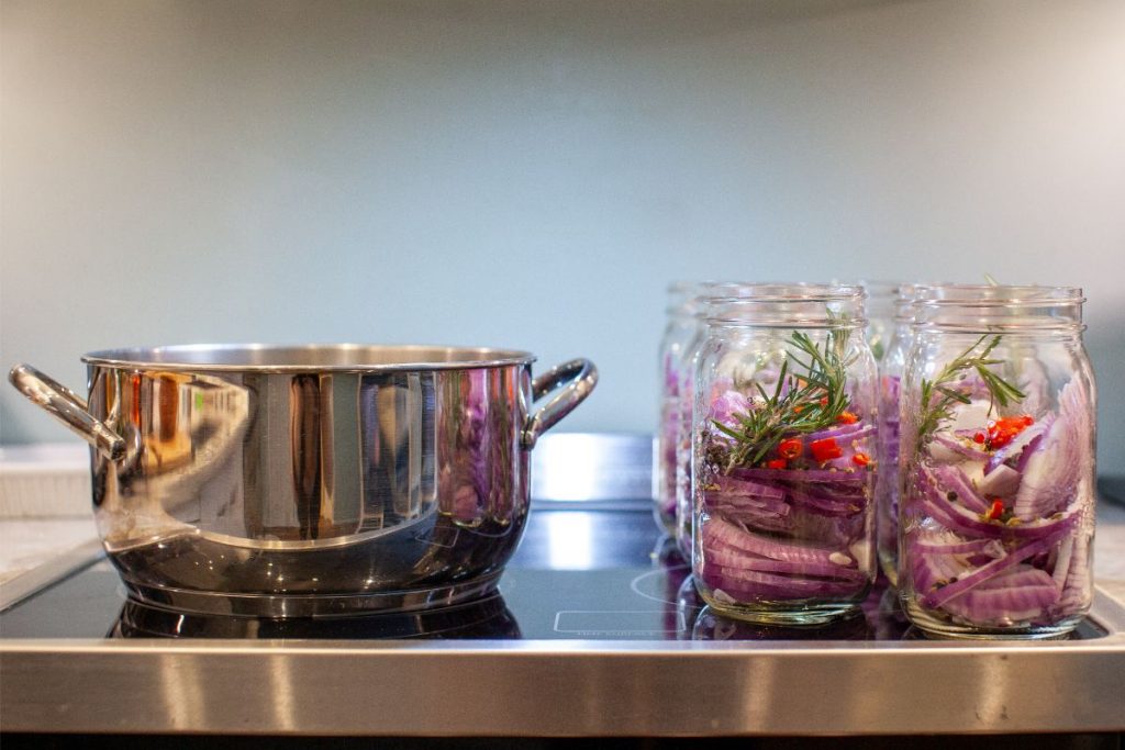 Jars of sliced red onions and pickling spices in canning jars on stove next to a pot of pickling brine