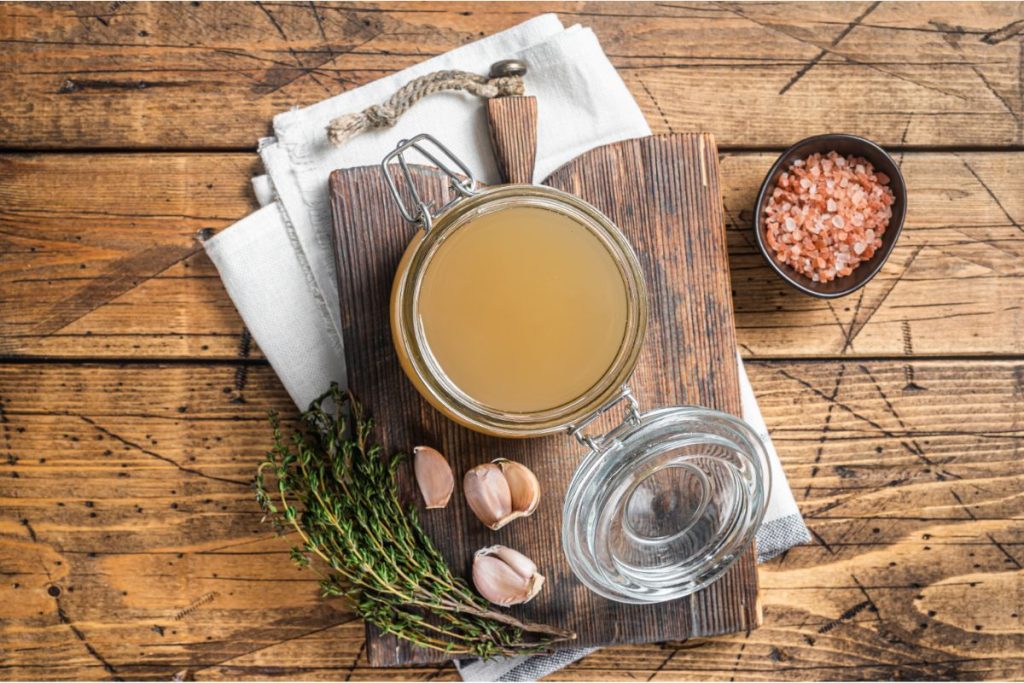 Open jar of bone broth on cutting board with raw garlic cloves, thyme, and Himalayan pink salt