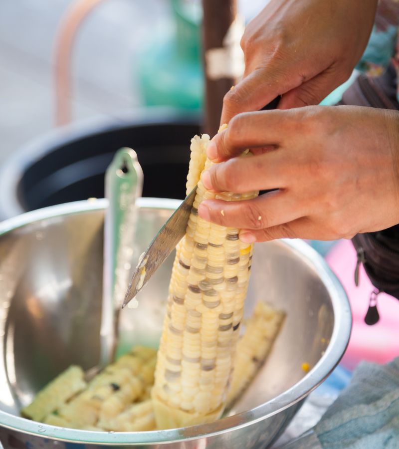Woman slicing corn kernels off the cob with a knife into a metal bowl