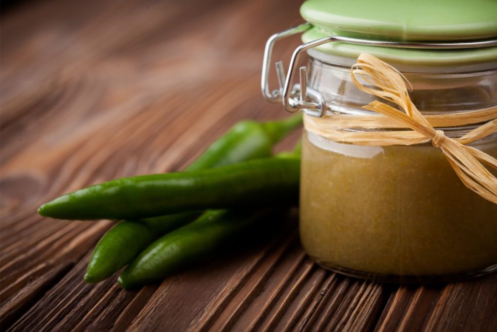 Green enchilada sauce in a small glass jar next to jalapeno peppers