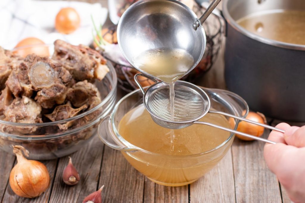 Pouring beef broth through a metal strainer to remove bones and fat