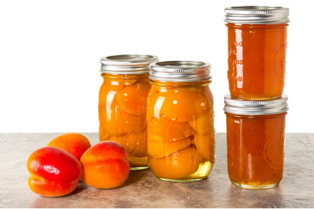 Jars of canned peaches and peach jam next to fresh peaches