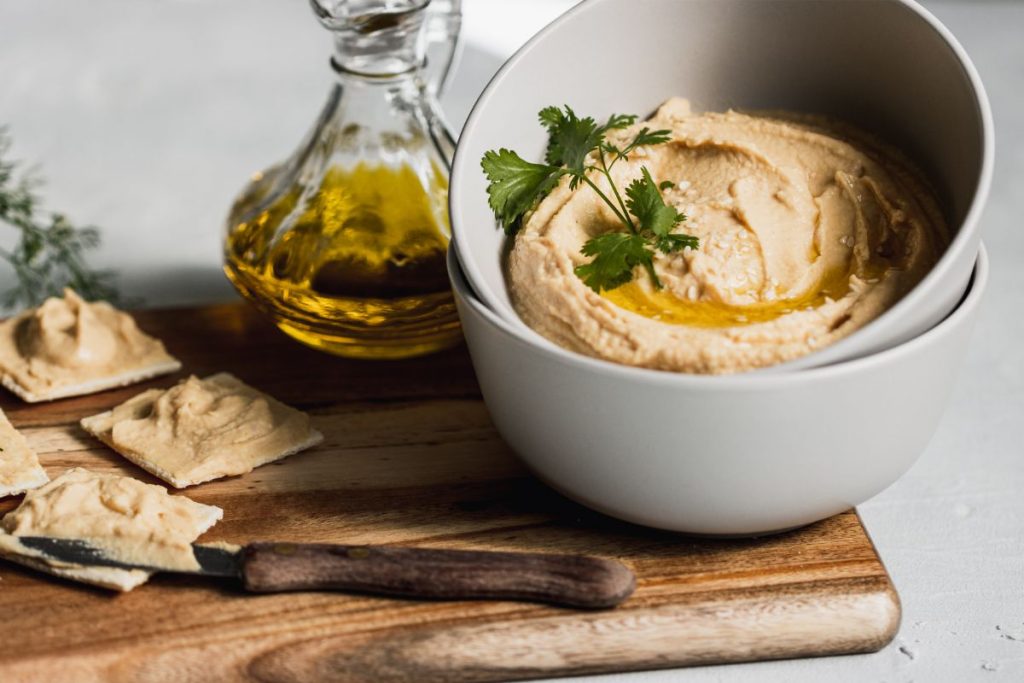 Bowl of prepared hummus with crackers and oil on a cutting board