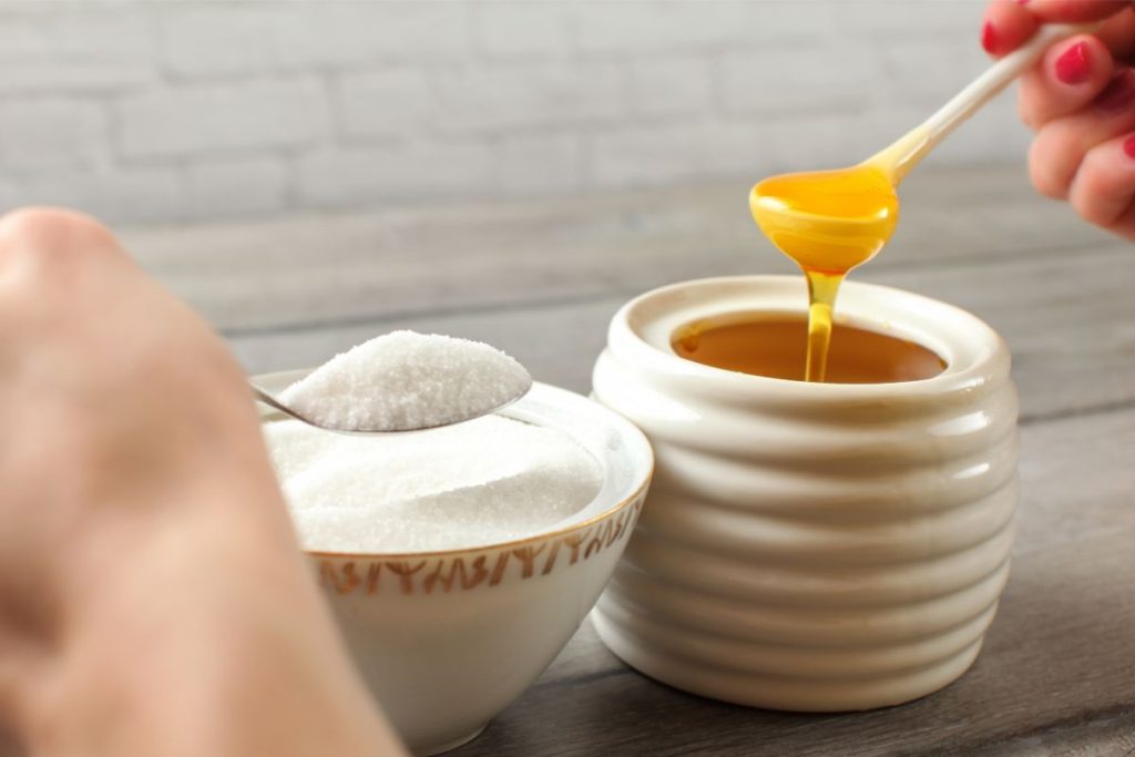 White sugar and honey syrup being lifted on spoons from two separate white containers