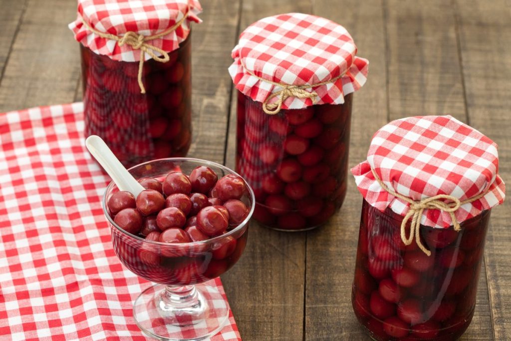 Jars of tart cherries canned in their own juice next to a bowl of tart cherries with a spoon