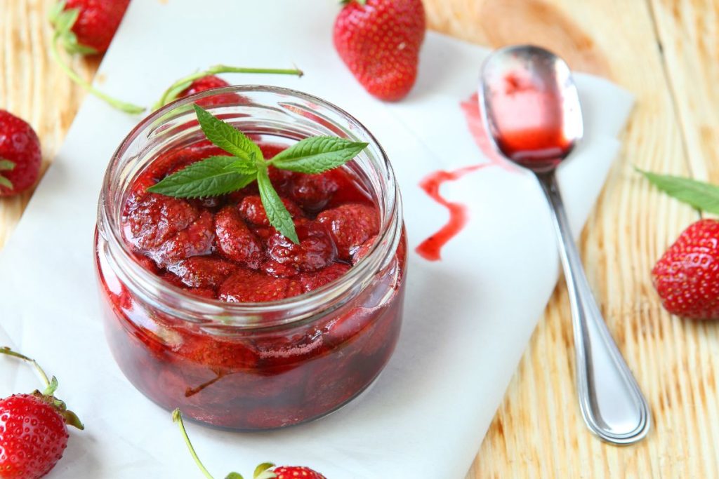 Whole strawberries canned and sitting in open canning jar with spoon next to the jar